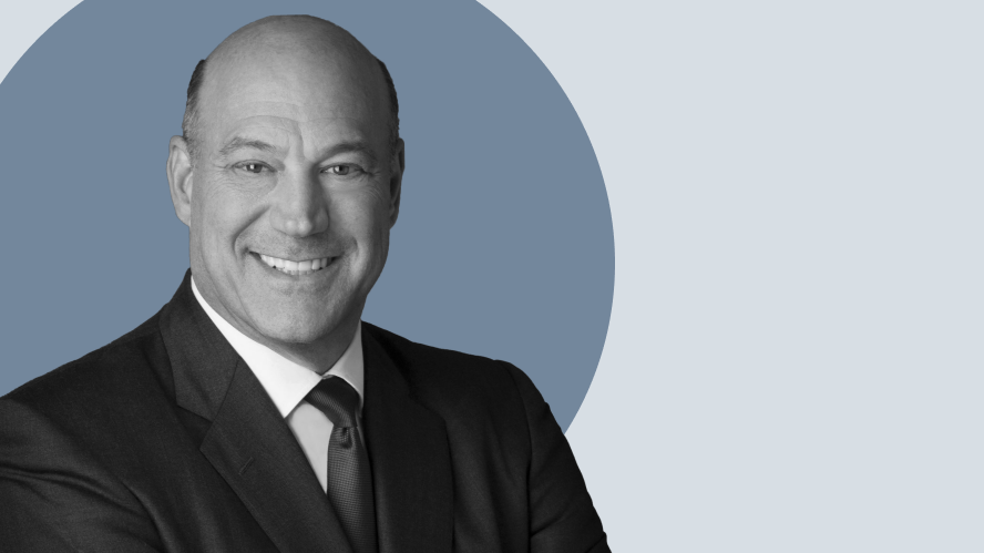 Chân dung của Gary Cohn, người đàn ông hói đầu trong bộ vest tối màu và đeo cà vạt, mỉm cười khoanh tay trước nền hình tròn màu xanh lam và xám nhạt.