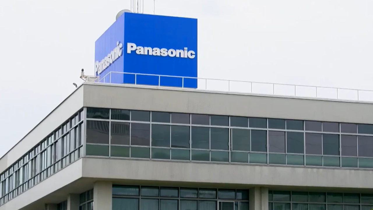 Tòa nhà thương mại hiện đại có cửa sổ kính và tấm biển Panasonic lớn màu xanh lam gắn trên sân thượng, đại diện cho Panasonic Holdings và bộ phận hệ thống ô tô của tập đoàn
